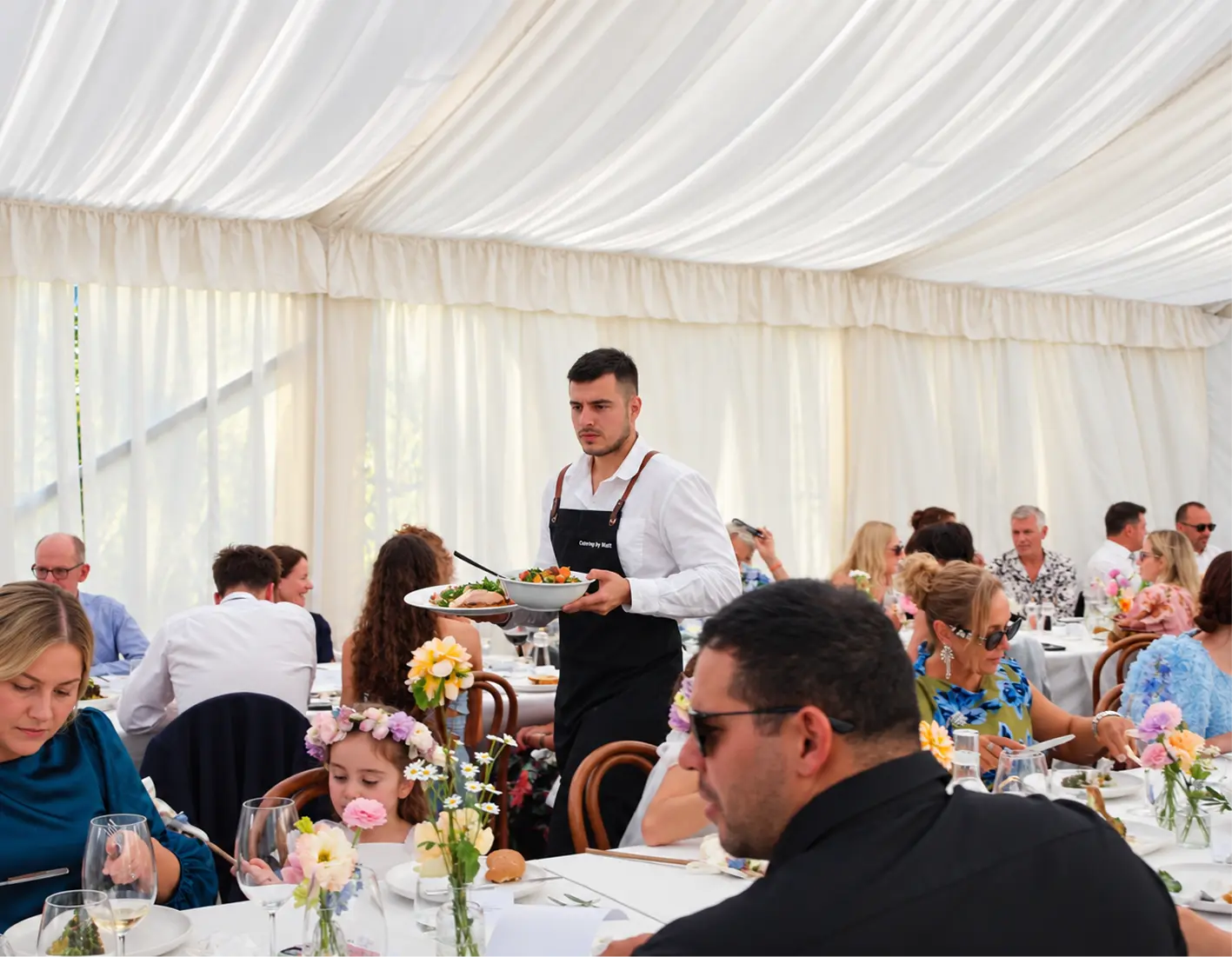 Waiter serving a shared dish to a full marquee of seated guests at a large home party