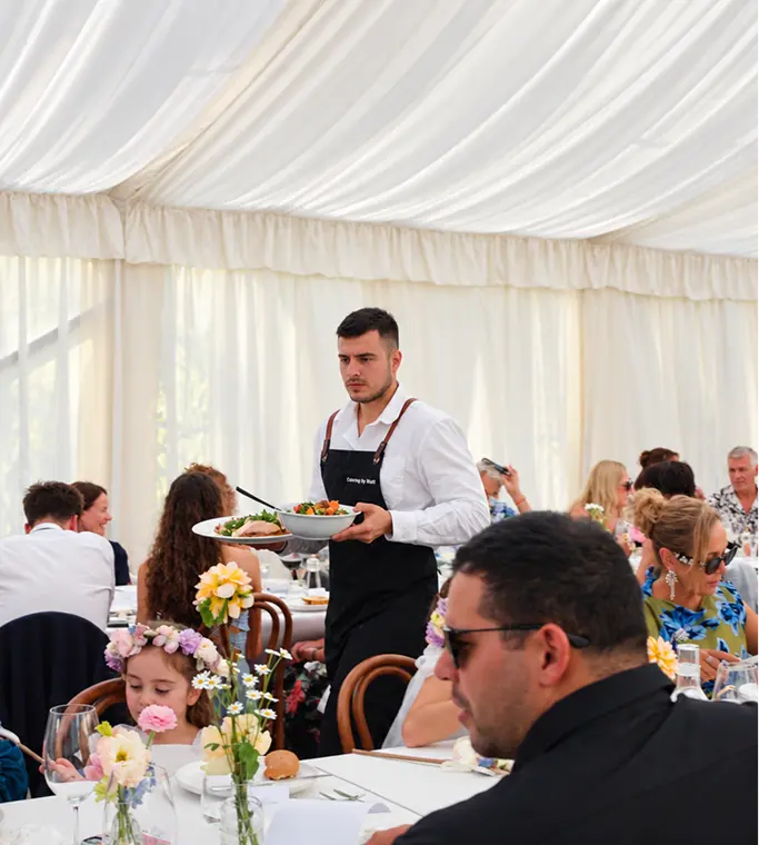 Waiter serving a shared dish to seated guests at a marquee celebration